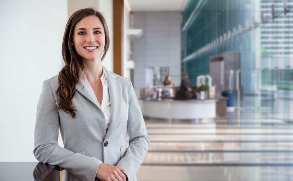 Business-Frau-Empfangsbereich Eine Frau mit Jackett steht angelehnt an einem Tisch in einer Empfangshalle und lächelt.