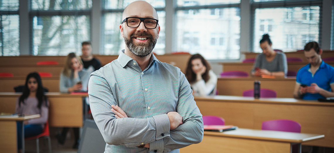 Ein Dozent steht mit verschränkten Armen im Vorlesungssaal und lächelt.