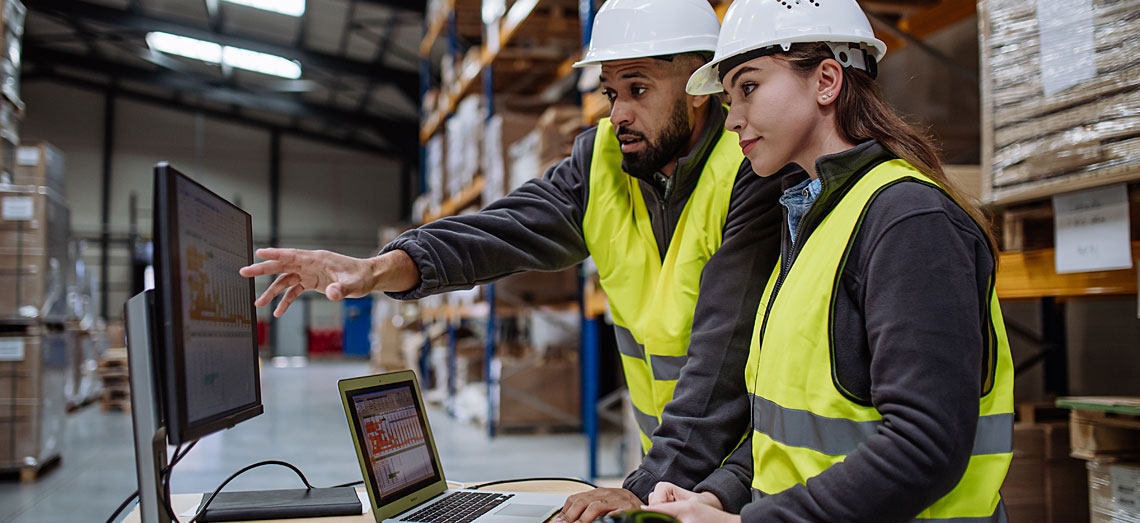 Fachwirt-fuer-Logistiksysteme-am-Laptop Ein Mann und eine Frau arbeiten im Lager an einem Laptop und vergleichen dort Zahlen.