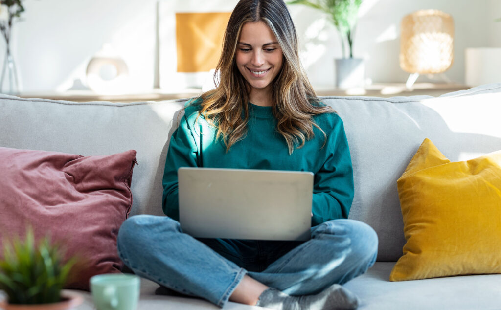Eine junge Frau sitzt im Schneidersitz auf einem Sofa und arbeitet mit einem Laptop.