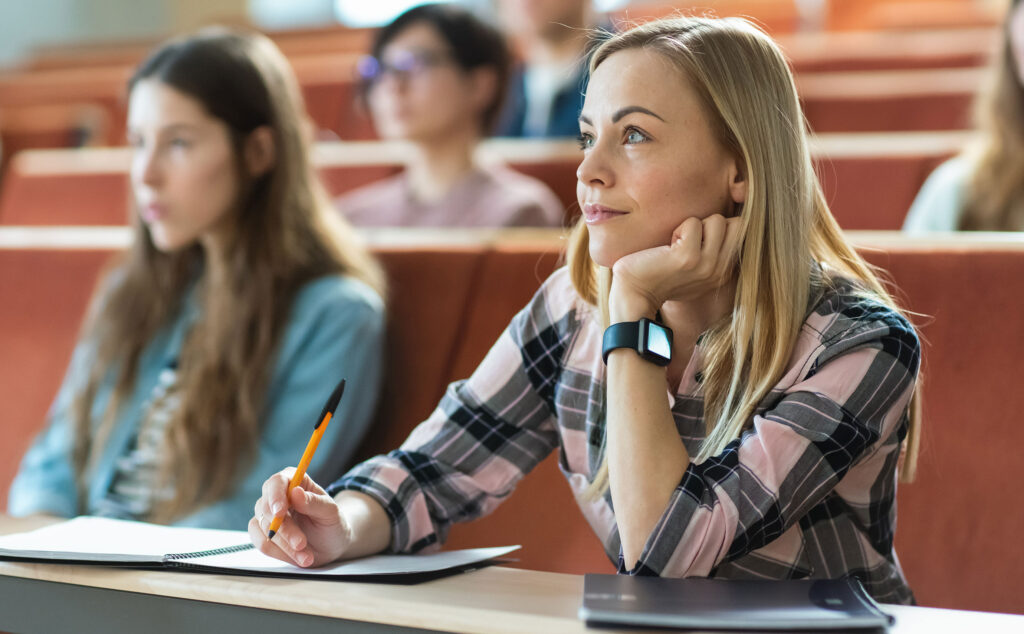 Eine Studentin sitzt mit einen Stift und Block im Hörsaal und schaut nach vorn.