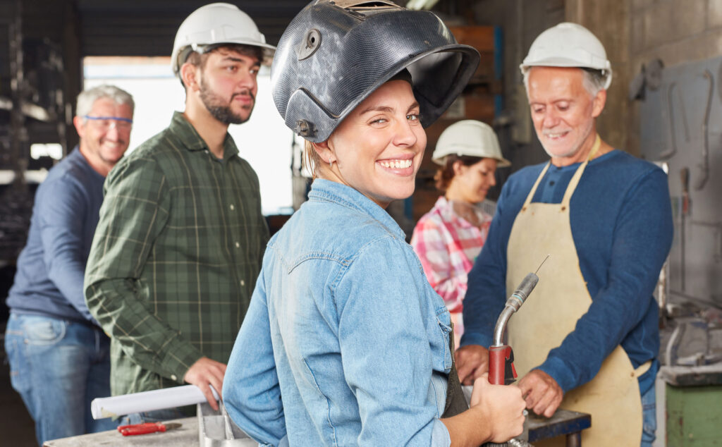 Eine Schlosserin steht mit einen Schweißgerät vor einem Arbeitstisch mit anderen Kollegen.
