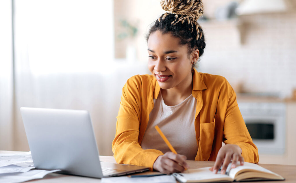 Eine Studentin sitzt vor dem Laptop und macht sich Notizen auf dem Block.
