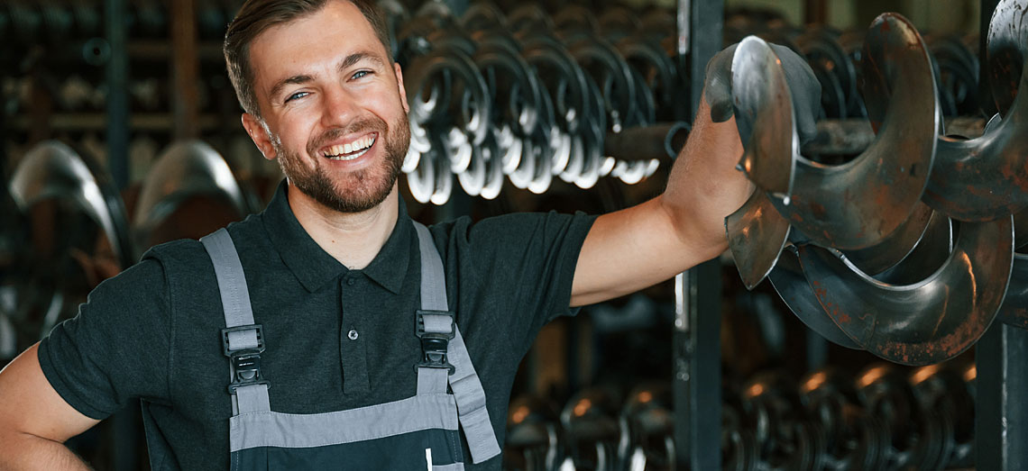 Ein Mann steht vor einem Lager mit Maschinenteilen und lächelt.