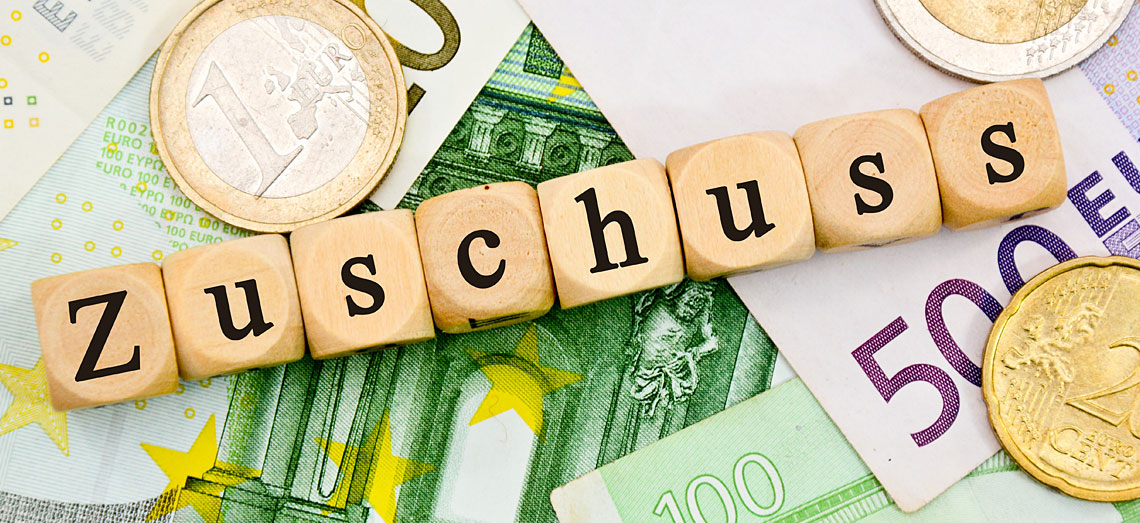 Holzwürfel mit dem Wort Zuschuss liegen auf Geldscheinen und Geldmünzen.