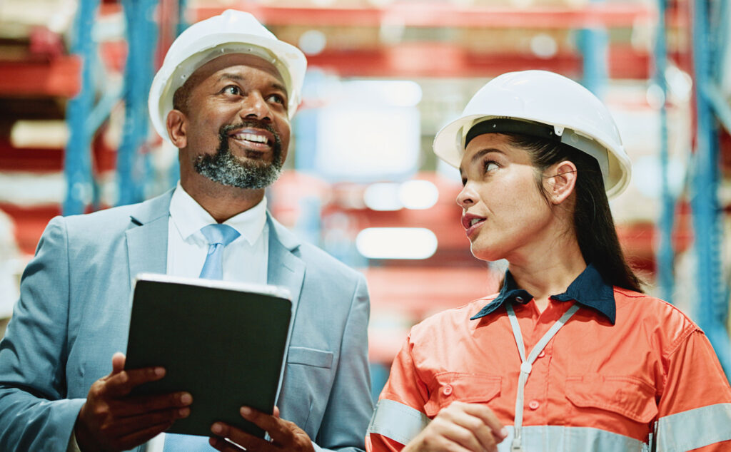 Eine Logistikmeisterin erklärt einem Besucher etwas in einer großen Lagerhalle.