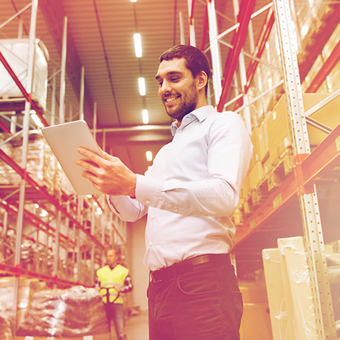 Ein Mann steht in einem Warenlager und schaut auf sein Tablet.