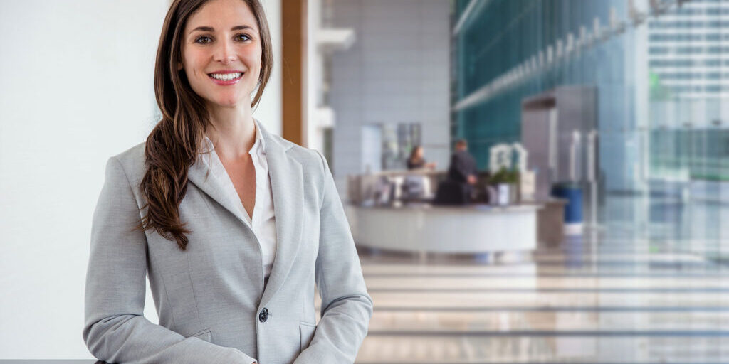 Eine Frau mit Jackett steht angelehnt an einem Tisch in einer Empfangshalle und lächelt.
