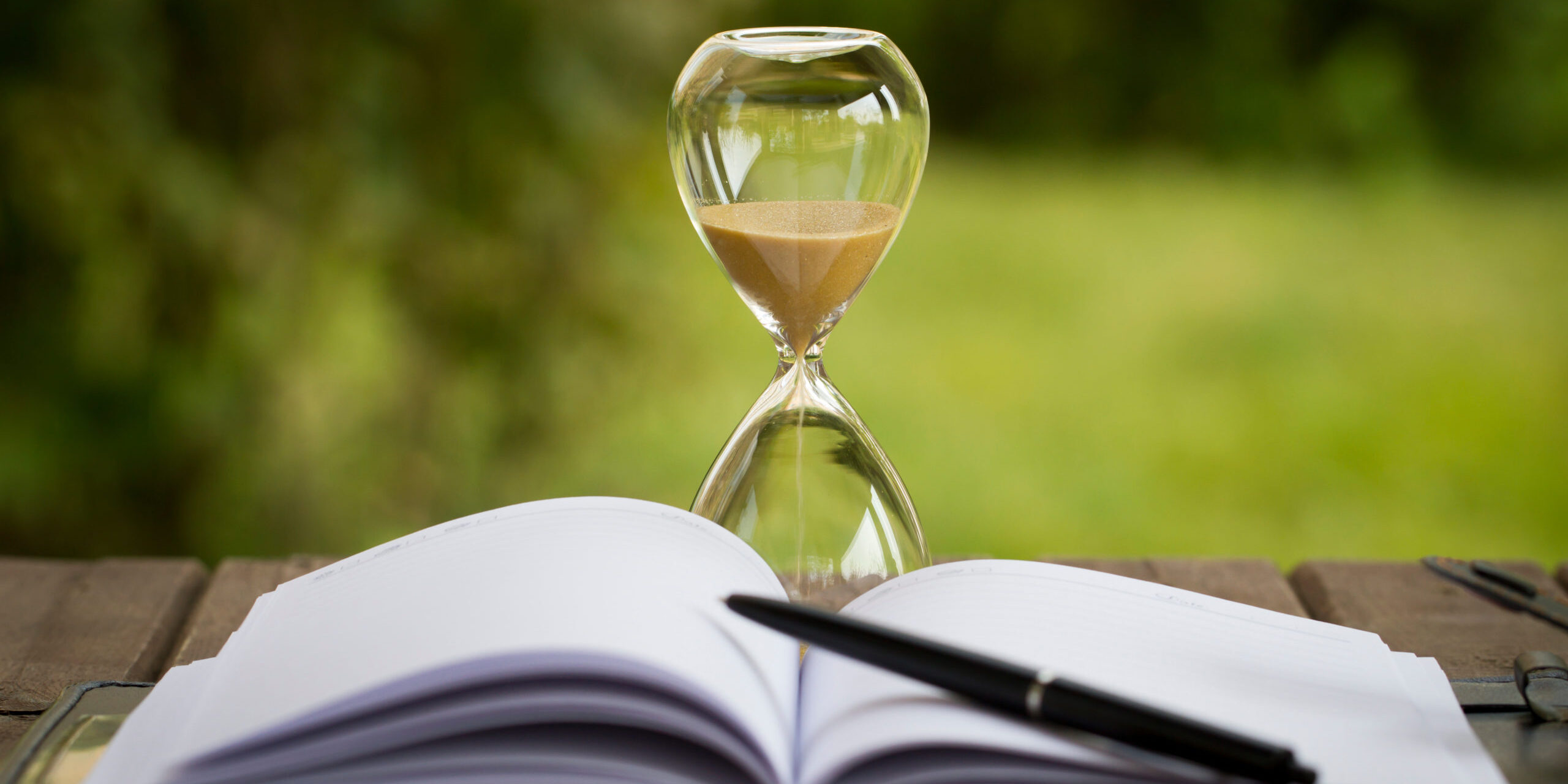 Eine Sanduhr steht hinter einem offenen Notizbuch, indem ein Stift liegt.